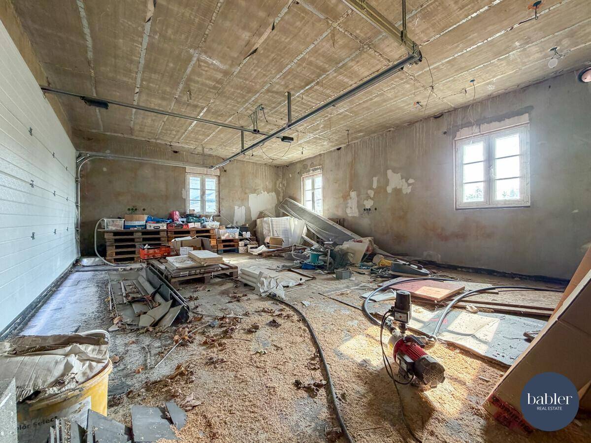 Garage im Rohbau mit Bauabfällen, sichtbaren Deckenbalken und Fenstern.