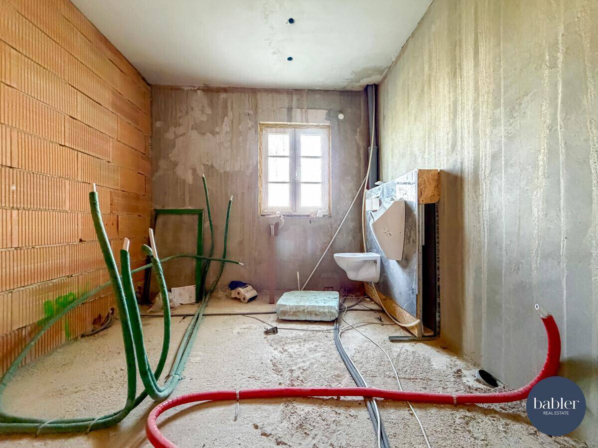 Badezimmer im Rohbau mit sichtbaren Installationen, Fenster und vorbereiteter Toilette.