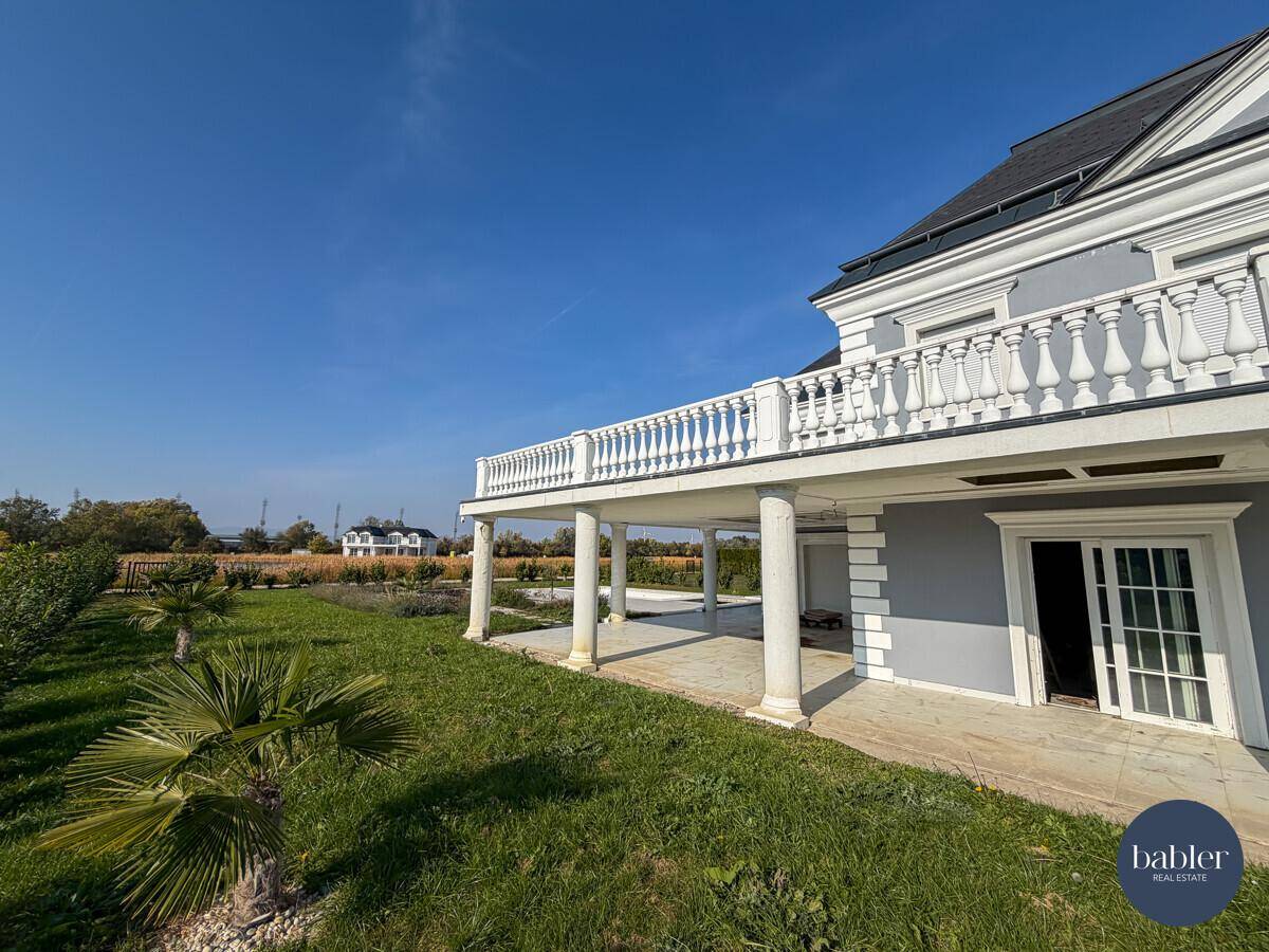 Seitlicher Blick auf die Villa mit überdachtem Terrassenbereich und junger Palmenbepflanzung.