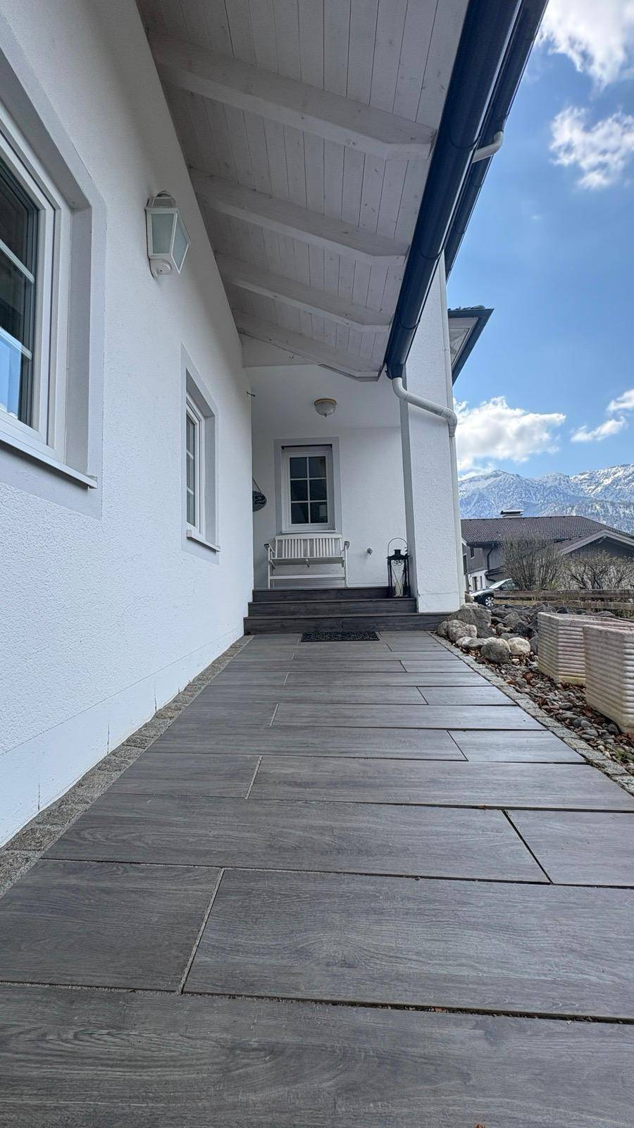 Langer, gefliester Weg entlang der Hausfassade mit Blick auf die Berge im Hintergrund.