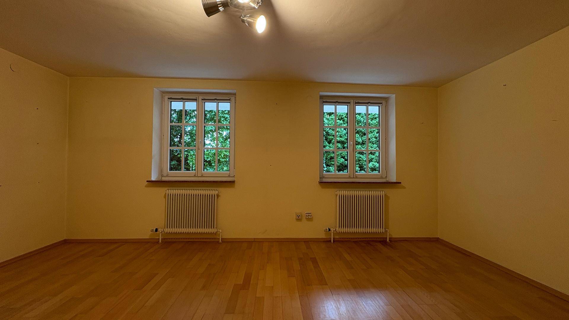 Heller, leerer Raum mit gelben Wänden, Holzboden und zwei großen Fenstern.