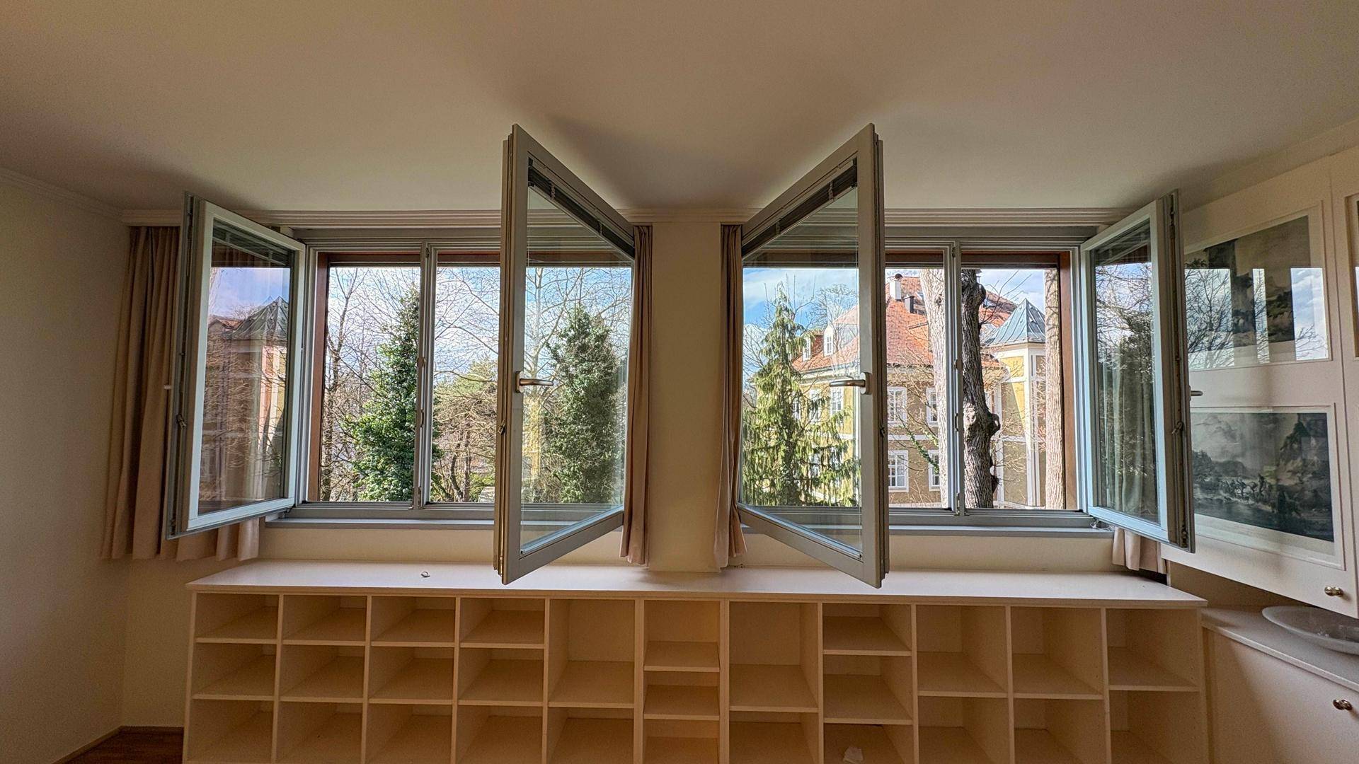 Heller Wohnbereich mit mehreren Fenstern, die einen grünen Ausblick bieten, und integrierten Regalen.