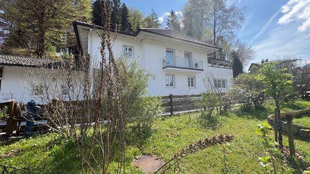Weißes Wohnhaus mit Balkon und Satteldach, umgeben von einem grünen Garten unter blauem Himmel.