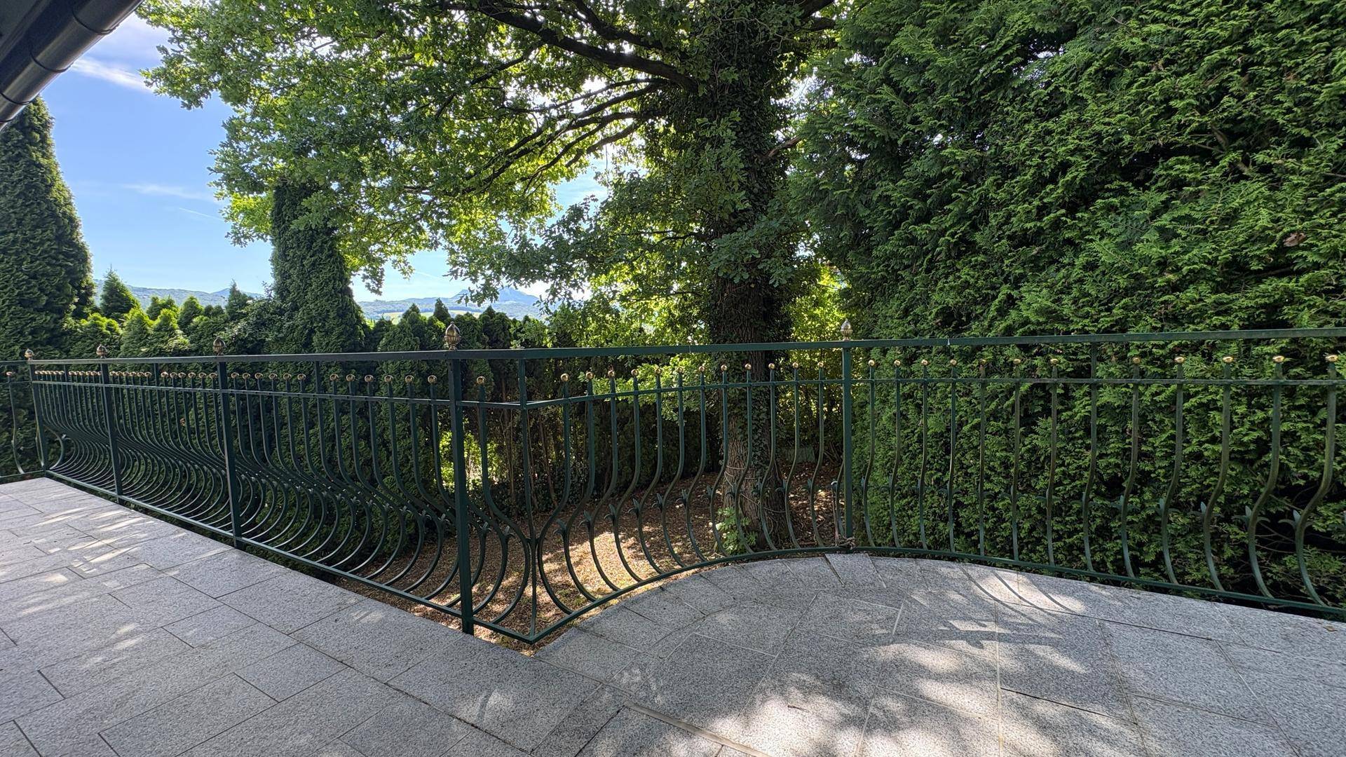 Geräumiger Balkon mit Steinboden und schmiedeeisernem Geländer, umgeben von Bäumen.