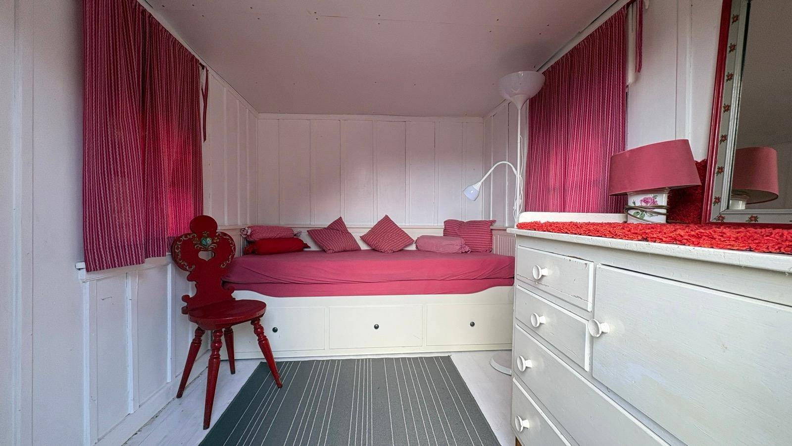 Kleines Schlafzimmer mit Tagesbett, weißen Holzpaneelen und roten Vorhängen.