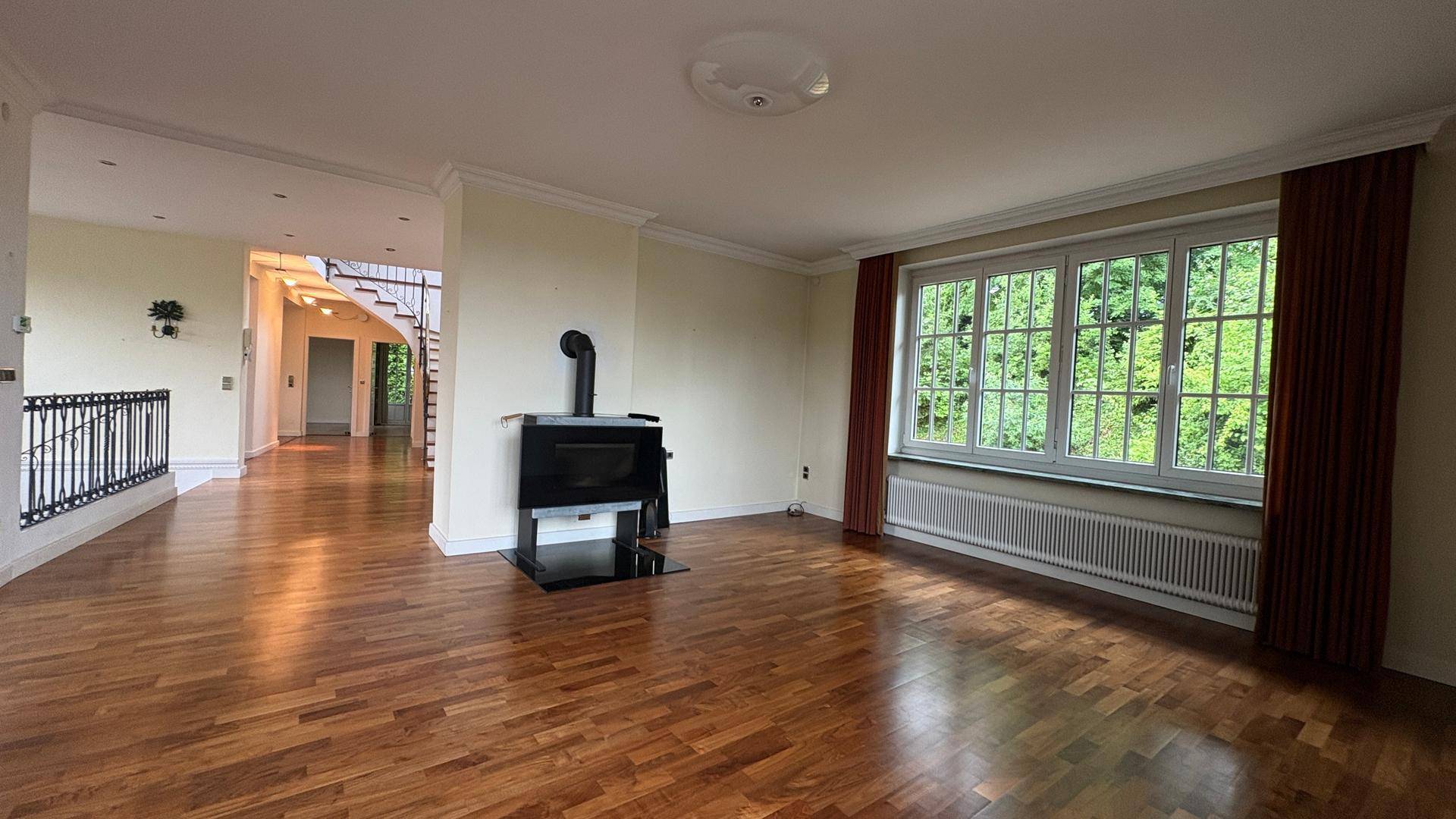 Geräumiges Wohnzimmer mit Holzboden, Kaminofen und großen Fenstern mit Grünblick.