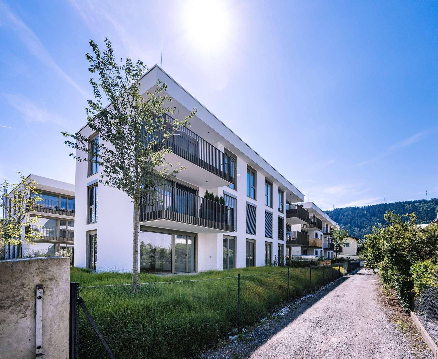 Neubau-Wohngebäude mit modernen Balkonen und gepflegter Grünfläche in alpiner Umgebung unter blauem Himmel.