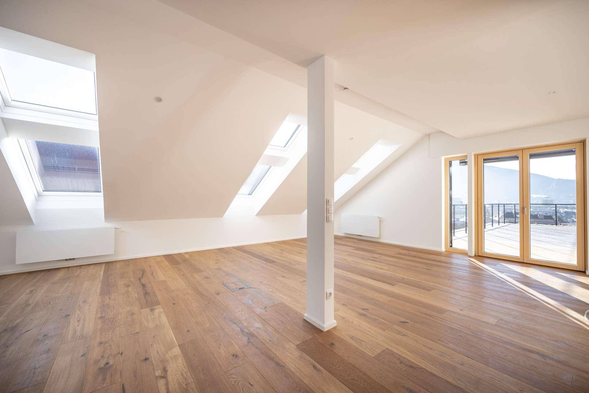 Heller Wohnraum mit Holzboden, Dachschrägen, mehreren Dachfenstern und Zugang zu einer Terrasse.