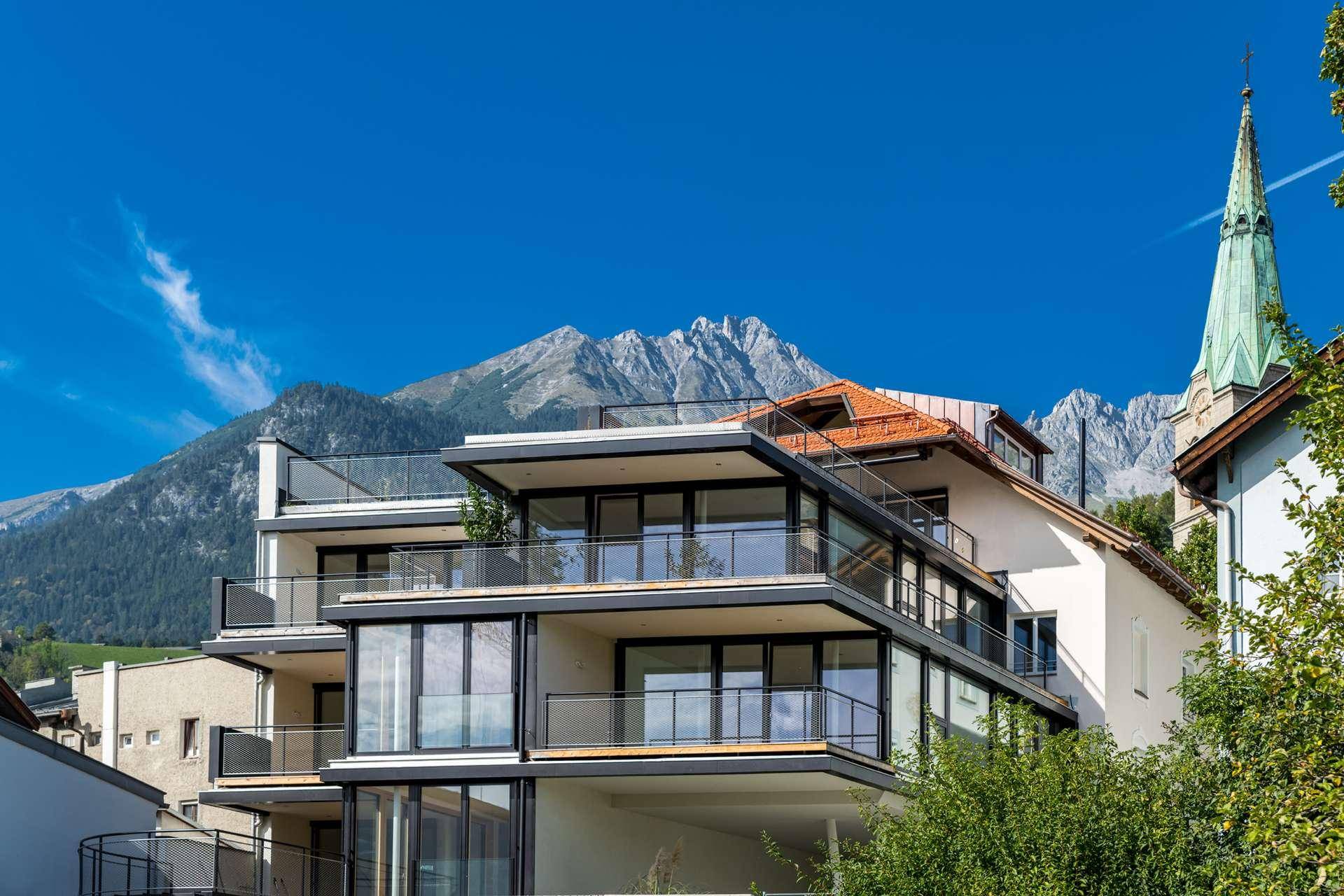 Moderne Architektur mit gestuften Balkonen und großen Glasflächen, eingebettet in eine Berglandschaft mit Kirchturm.