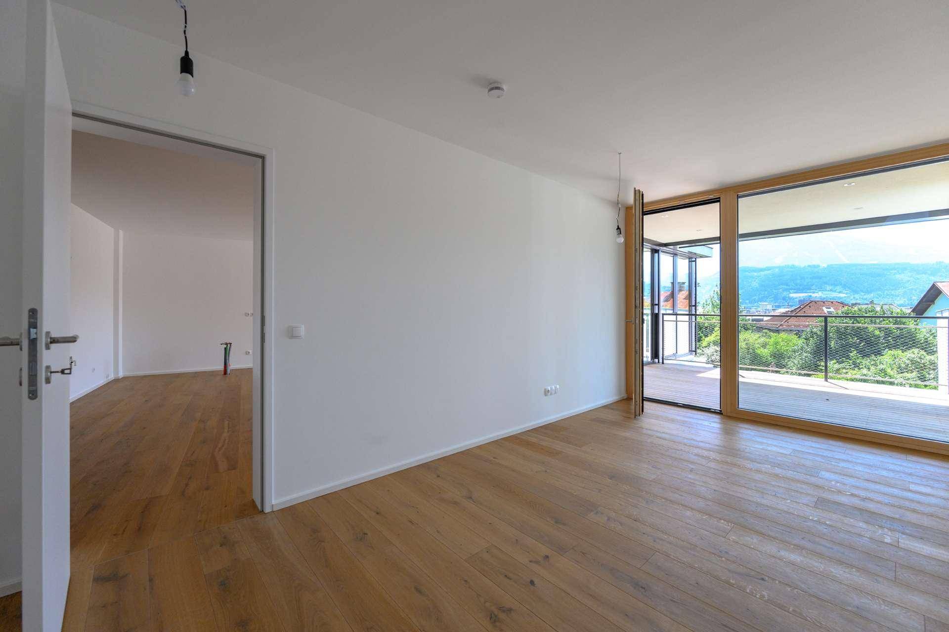 Heller Wohnbereich mit Holzboden und raumhohen Fenstern, die Zugang zu einem Balkon mit Bergblick ermöglichen.