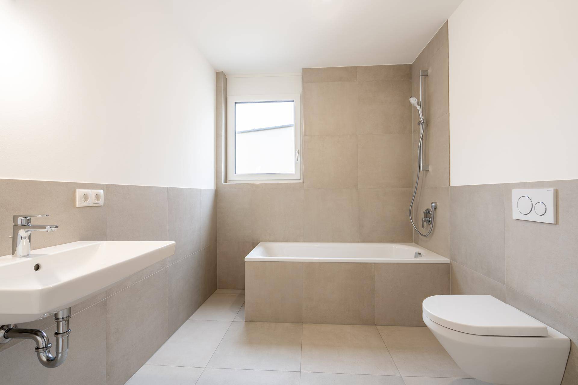 Modernes Badezimmer mit beigefarbenen Fliesen, Badewanne, wandhängendem WC und Fenster.