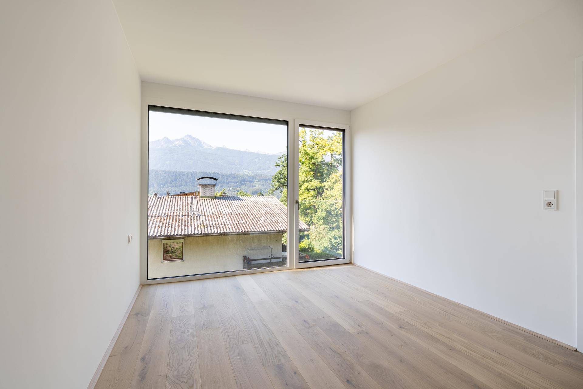 Helles Zimmer mit Holzboden und Fenster, das einen Blick auf ein Dach und Berge bietet.