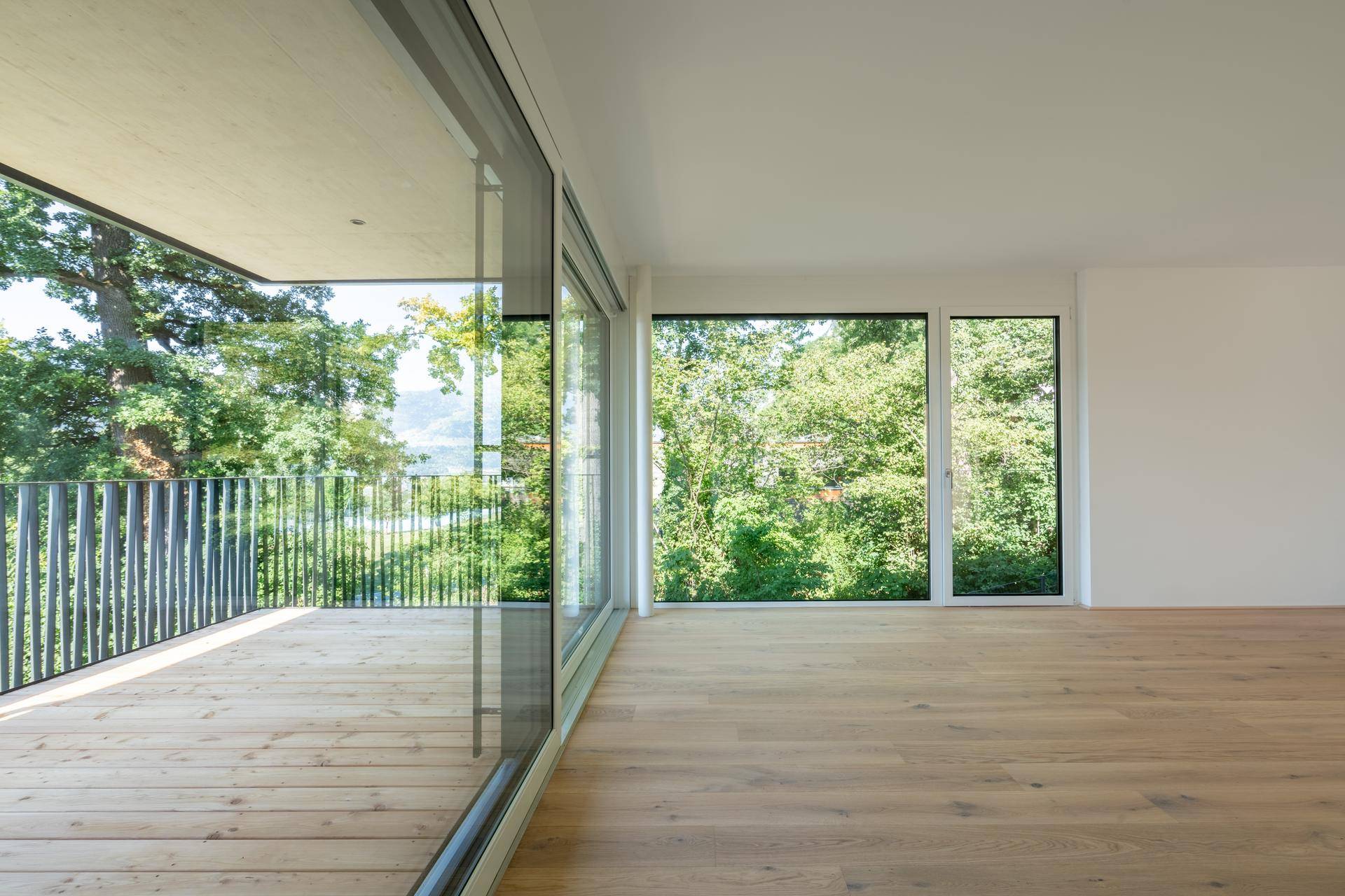 Lichtdurchfluteter Raum mit Holzboden und großem Balkon, umgeben von grünen Bäumen.