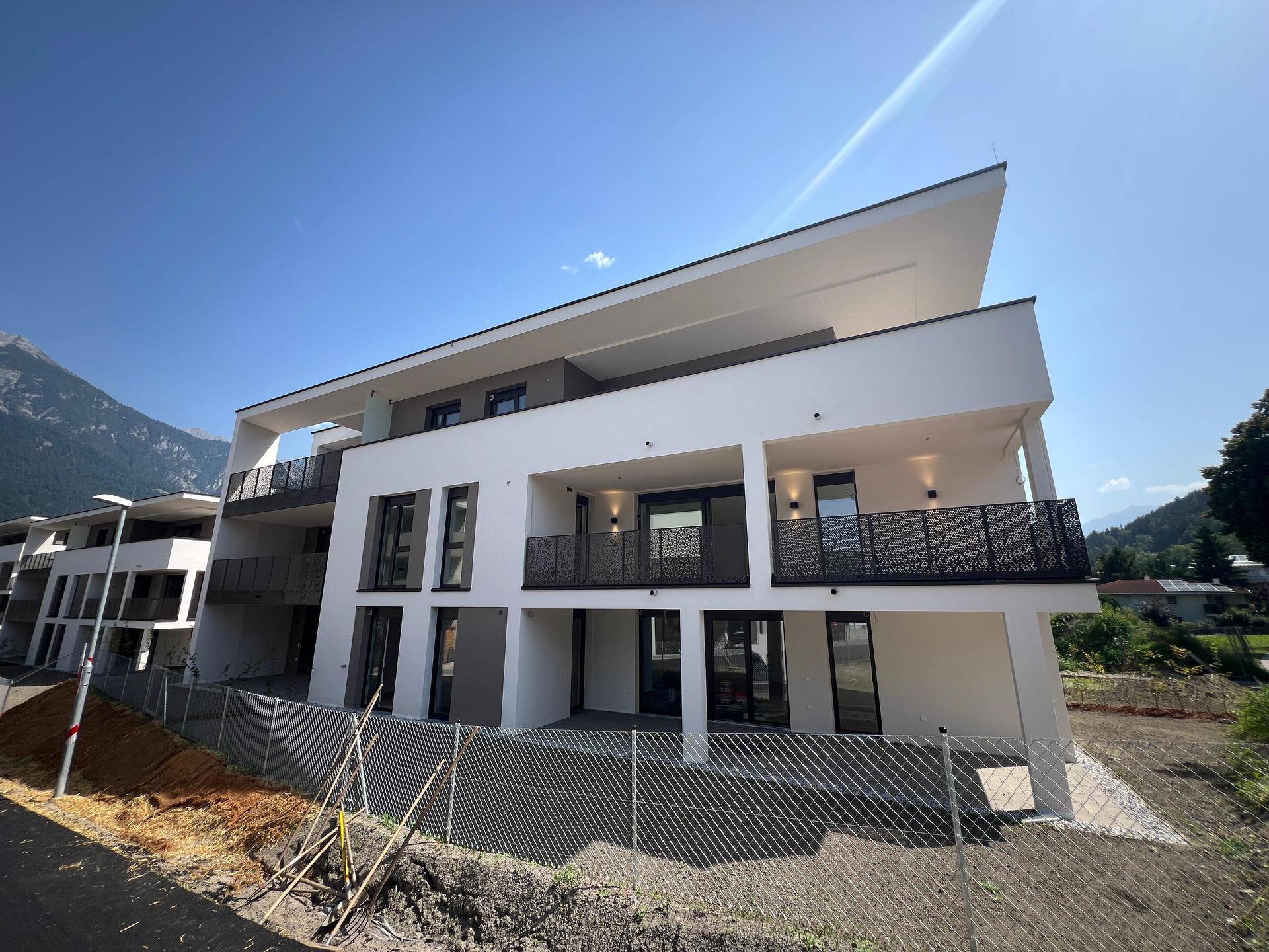 Neubau mit moderner Architektur, großen Fenstern und Balkonen in bergiger Umgebung bei Sonnenschein.