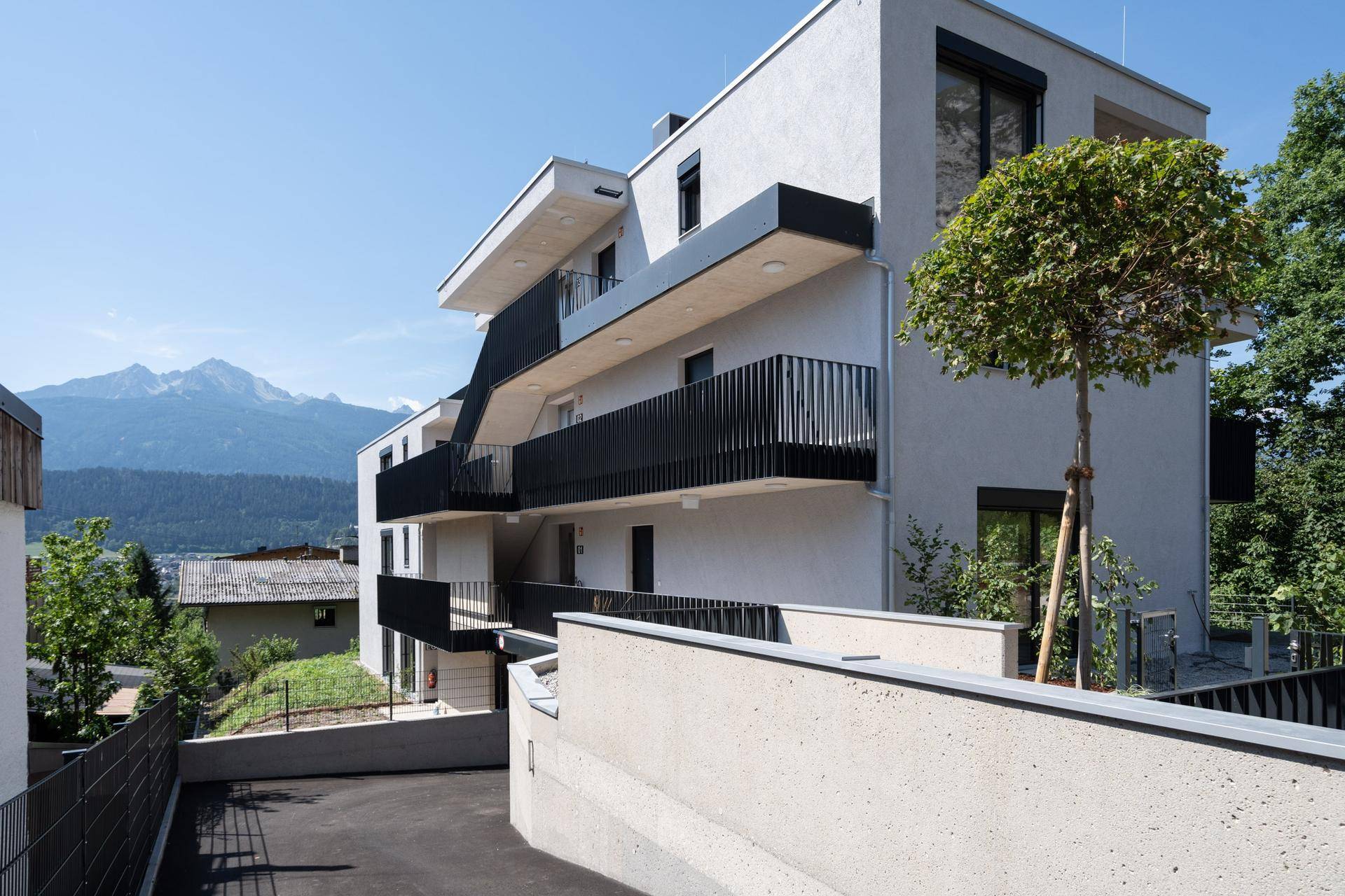 Neubau mit moderner Architektur, Balkonen und Zufahrt, umgeben von Natur und Bergen.