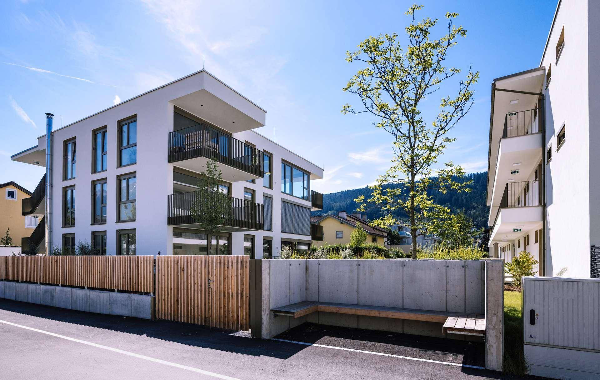Moderne Wohnanlage mit Balkonen, Holzzaun und integrierter Sitzbank im Außenbereich vor Bergkulisse.