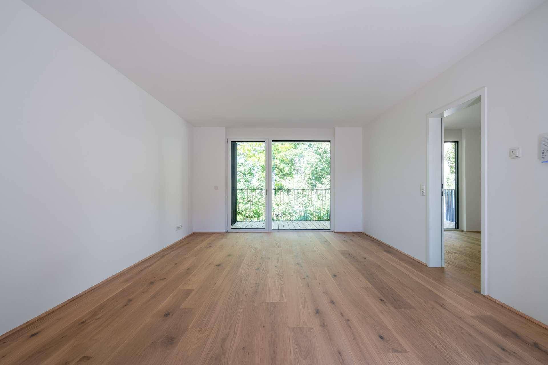 Heller, leerer Wohnraum mit Eichenparkett und großem Fenster, das Zugang zu einem Balkon mit Grünblick bietet.