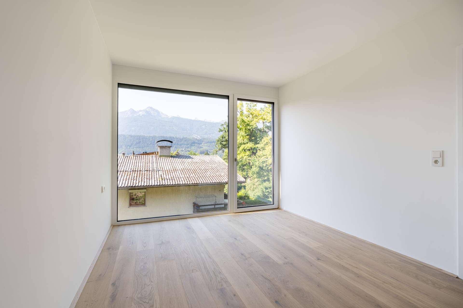 Heller Raum mit Holzboden und großem Fenster, das einen Ausblick auf die umliegende Landschaft und Berge bietet.