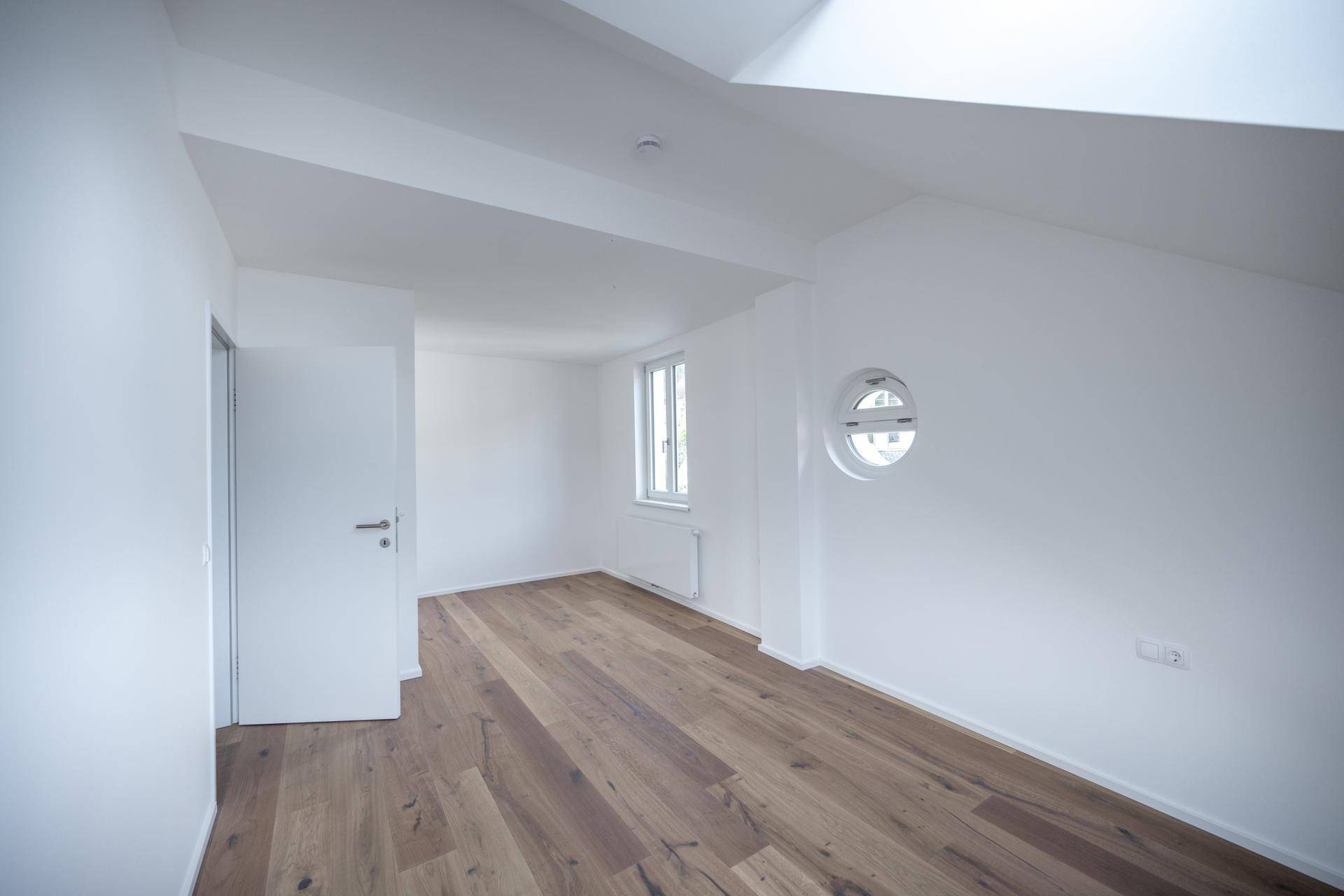 Lichtdurchfluteter Raum mit Holzboden, runden und rechteckigen Fenstern, ideal als Schlafzimmer.
