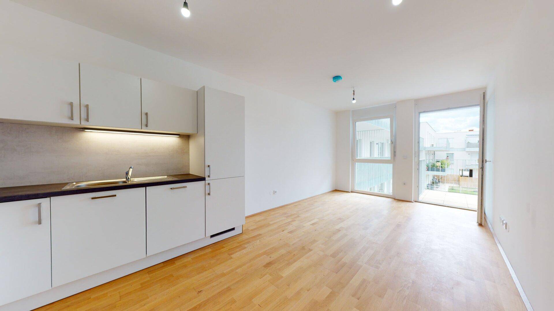 Heller offener Wohnbereich mit Holzboden, Zugang zum Balkon und integrierter Küchenzeile.
