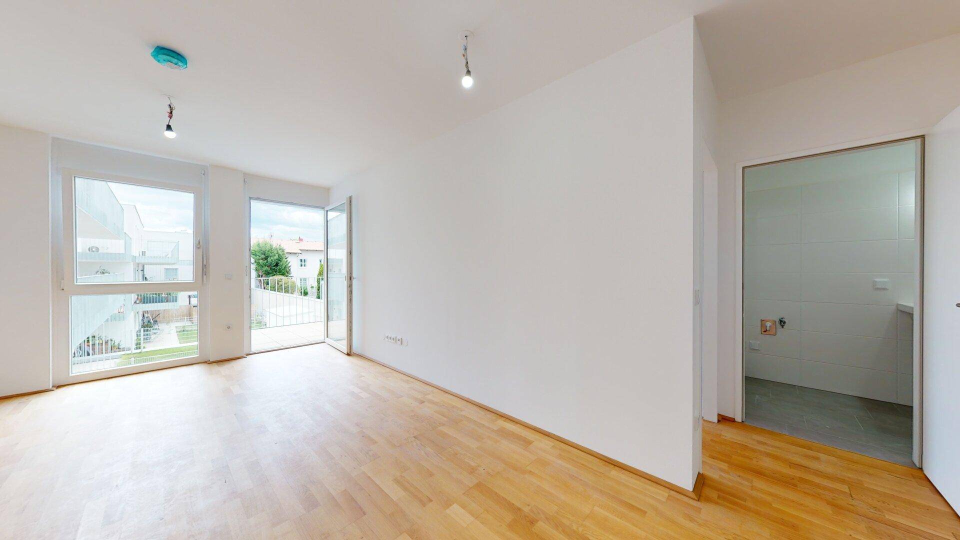 Heller Raum mit Holzboden, Zugang zum Balkon und Blick in ein gefliestes Badezimmer.