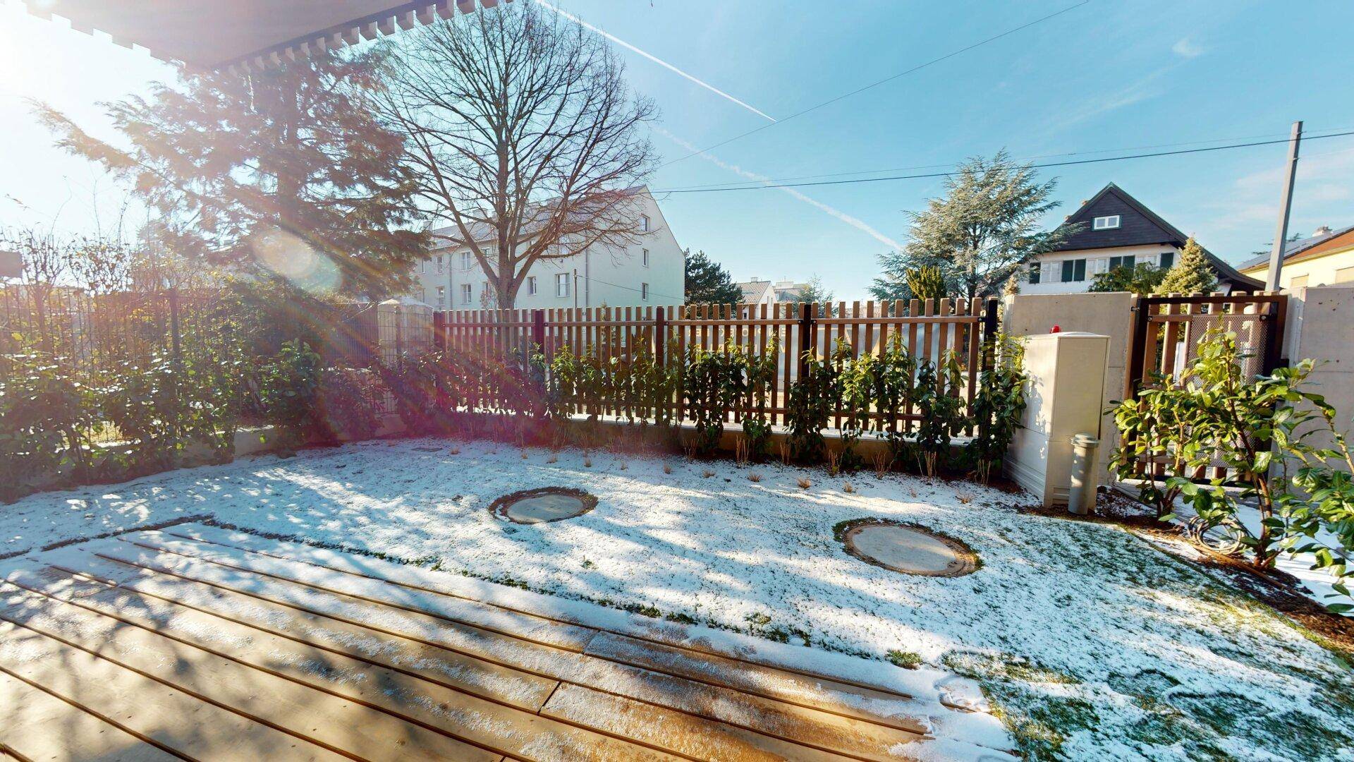 Gartenbereich mit Holzterrasse und Rasenfläche, leicht mit Schnee bedeckt, umgeben von einem Zaun.