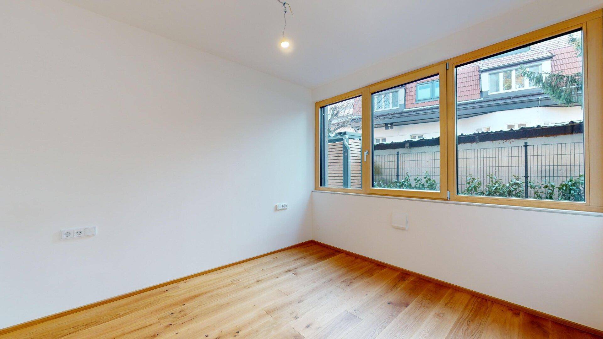 Helles Zimmer mit Holzboden und Fenster, das einen Blick auf die Nachbarschaft bietet.