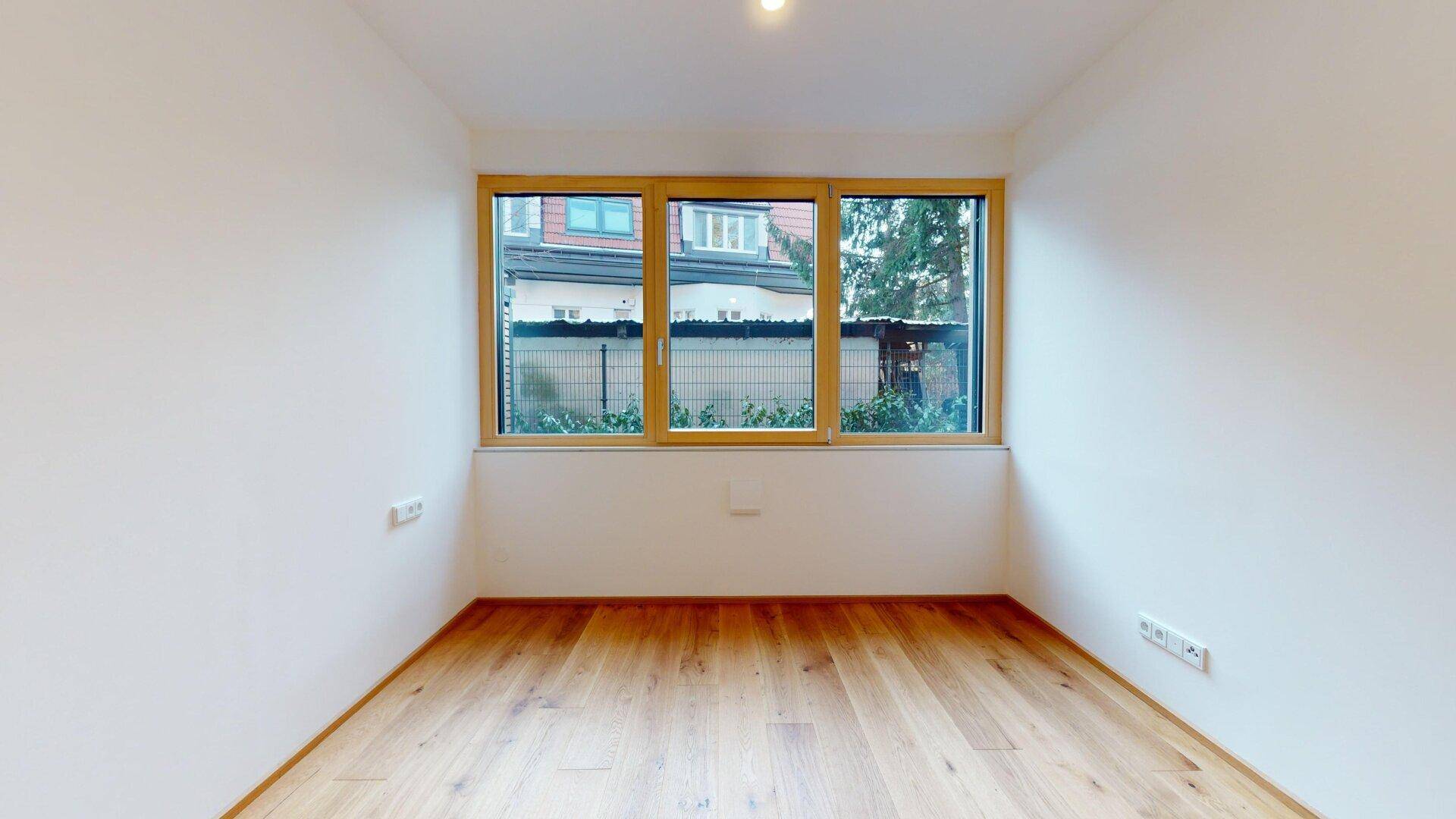 Leeres Zimmer mit Holzboden und großem Fenster, das viel Tageslicht hereinlässt.