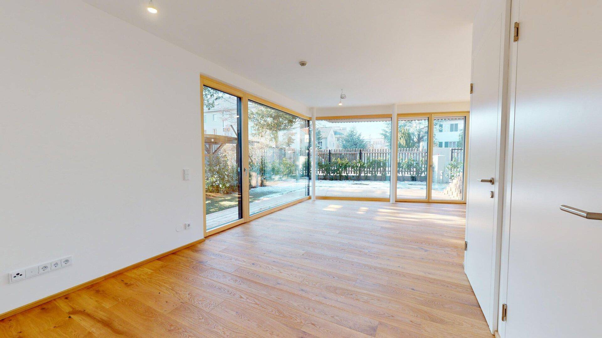 Geräumiges Wohnzimmer mit Holzboden und großen Fenstern, die viel Tageslicht hereinlassen.