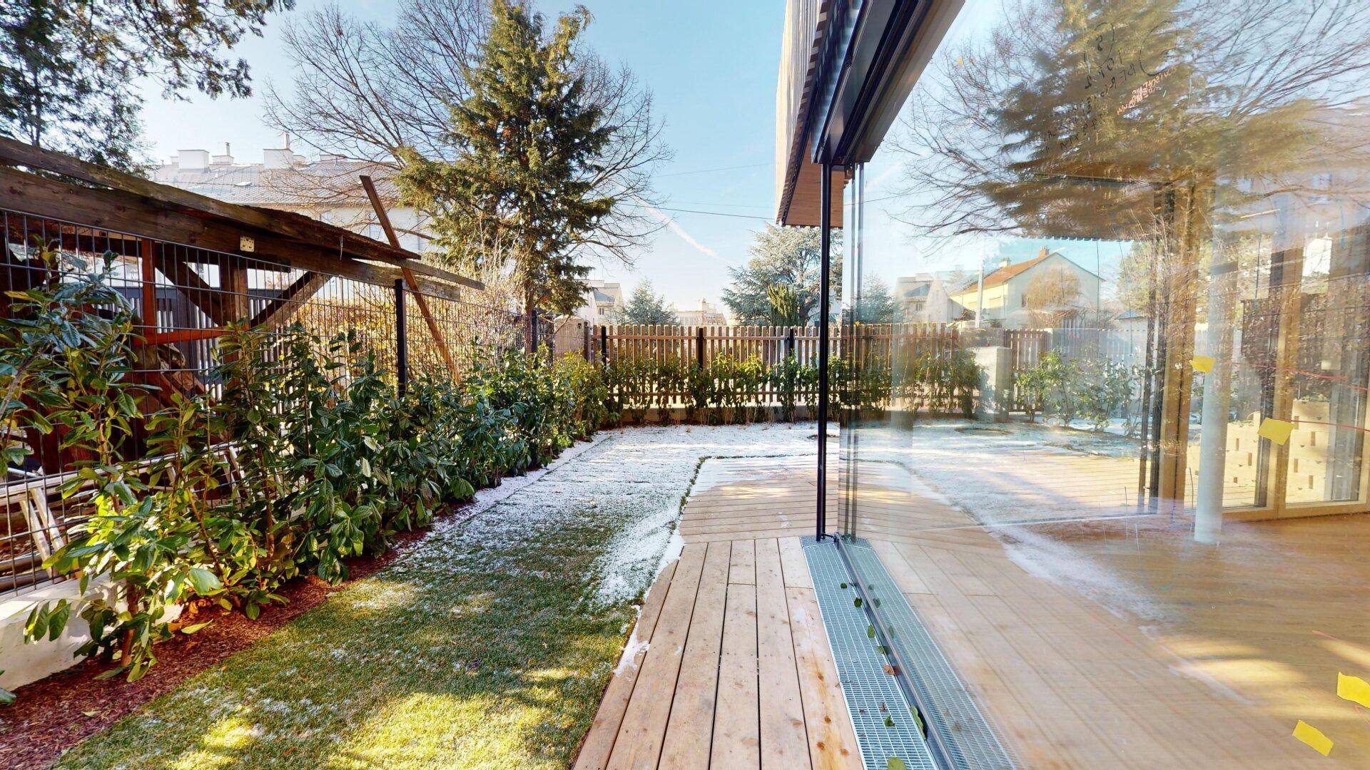 Seitlicher Gartenbereich mit Holzterrasse und Glasfassade des Gebäudes, umgeben von Bepflanzung.