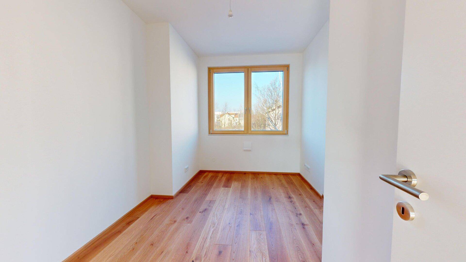 Kleines, helles Schlafzimmer mit Holzboden und einem Fenster, das einen ruhigen Blick ins Grüne bietet.