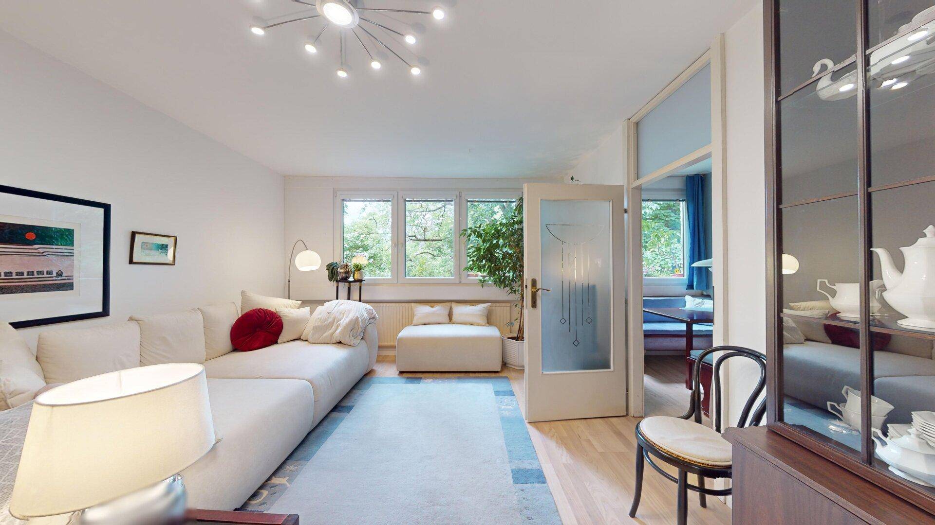 Geräumiges Wohnzimmer mit hellem Sofa, Teppich und Blick ins Grüne durch große Fenster.