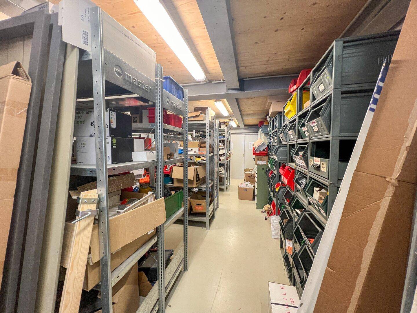 Schmaler Gang in einem Lagerraum mit Regalen voller Kleinteile und Kartons unter einer Holzdecke.