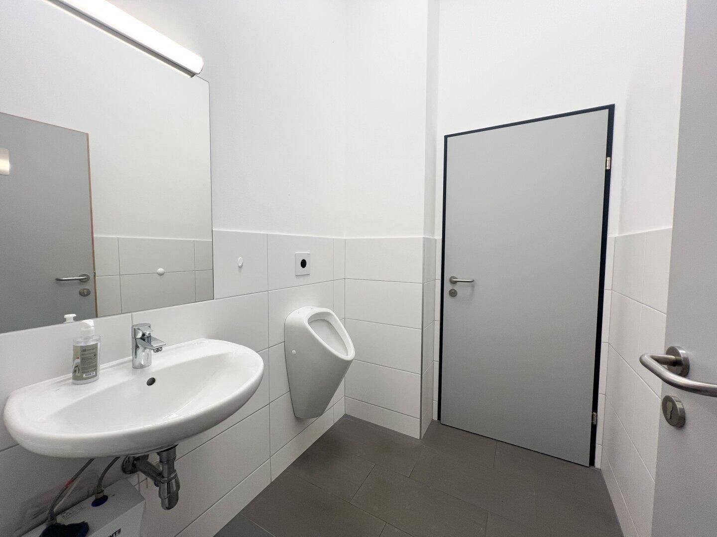 Saubere und moderne Toilette mit Urinal, Waschbecken und großen Fliesen in neutralen Farben.