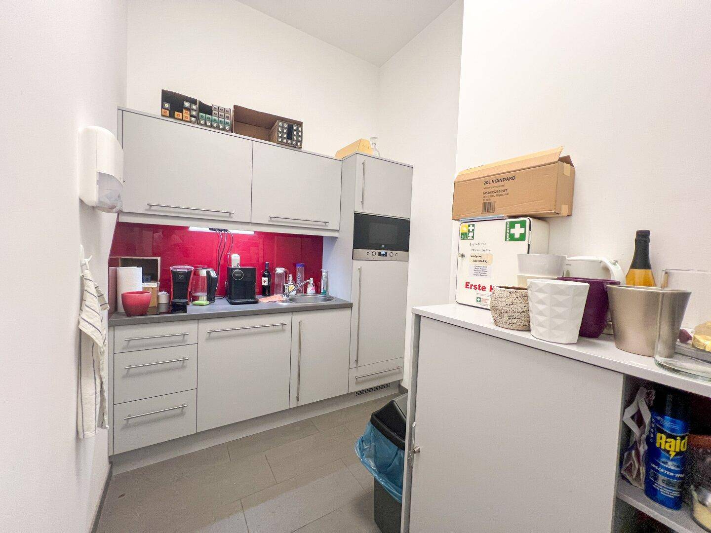 Kleine, moderne Küchenzeile mit grauen Schränken, roter Rückwand und integrierter Mikrowelle in einem Bürogebäude.