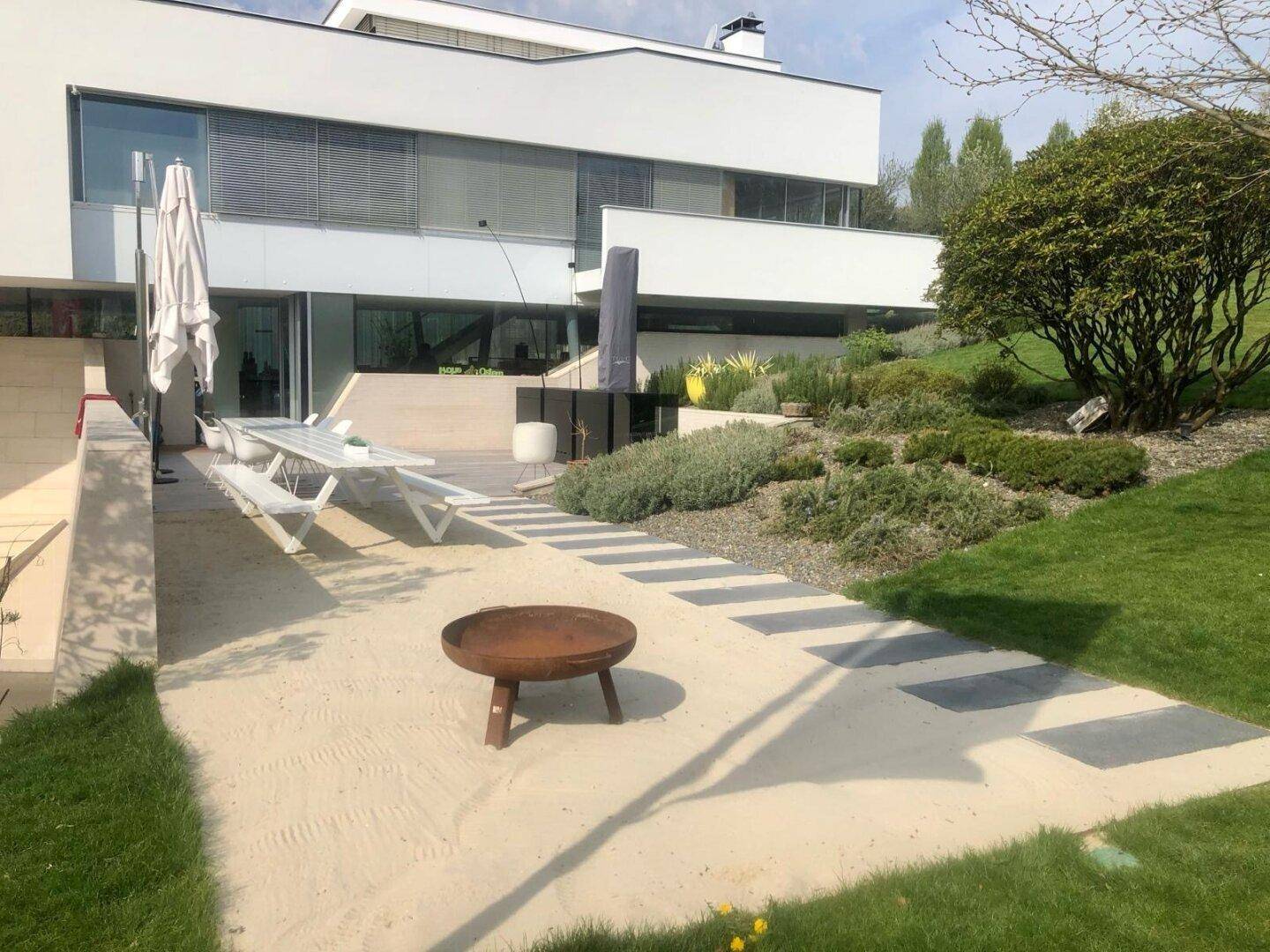 Großzügiger Gartenbereich mit Sandfläche, Feuerstelle und Sitzgelegenheiten vor der modernen Villa.