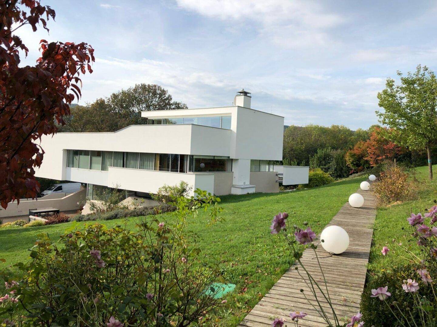 Moderne Villa in Hanglage, umgeben von einer weitläufigen Grünfläche mit Bäumen und Gartenwegen.