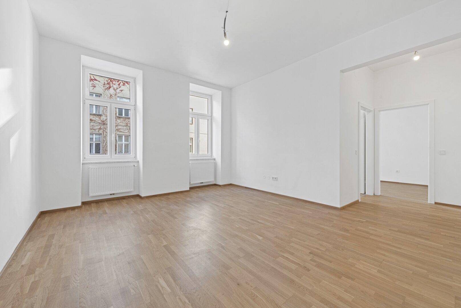 Helles Zimmer mit Parkettboden und zwei hohen Fenstern, ideal für Wohn- oder Schlafbereich.