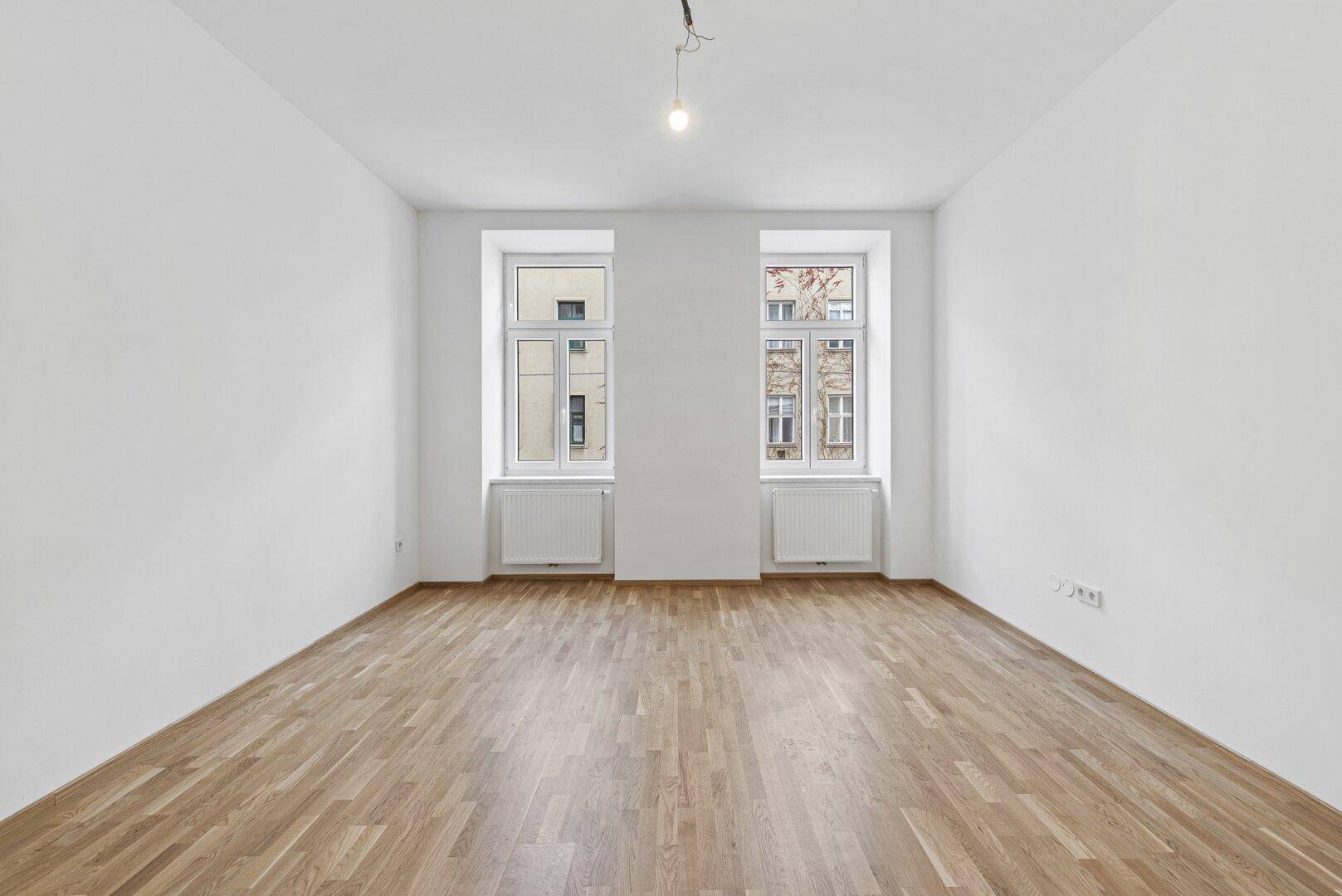 Heller, leerer Raum mit Parkettboden und zwei großen Fenstern, ideal für Wohnzwecke.