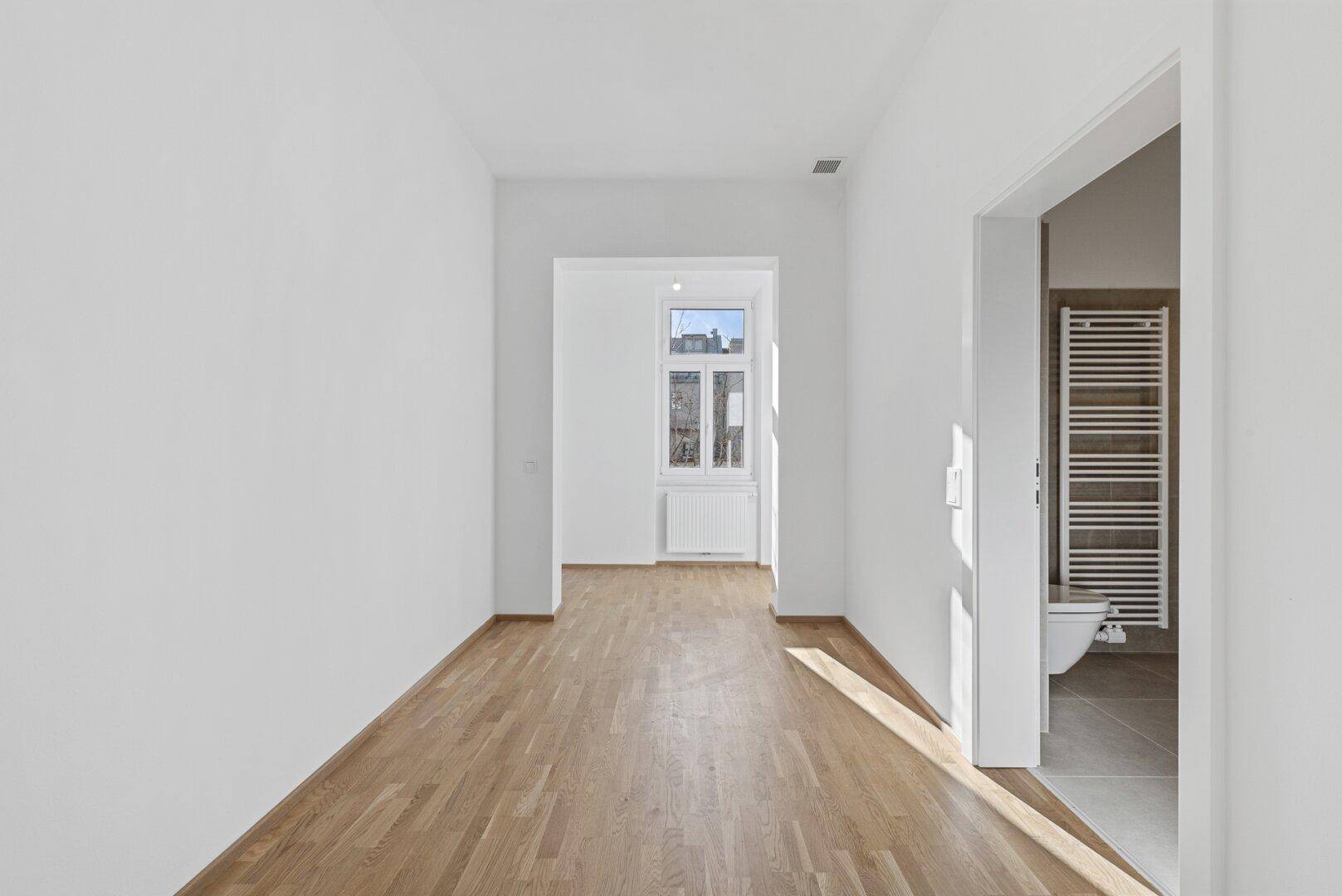 Heller Flur mit Parkettboden, Fenster und Zugang zu einem modernen Badezimmer.