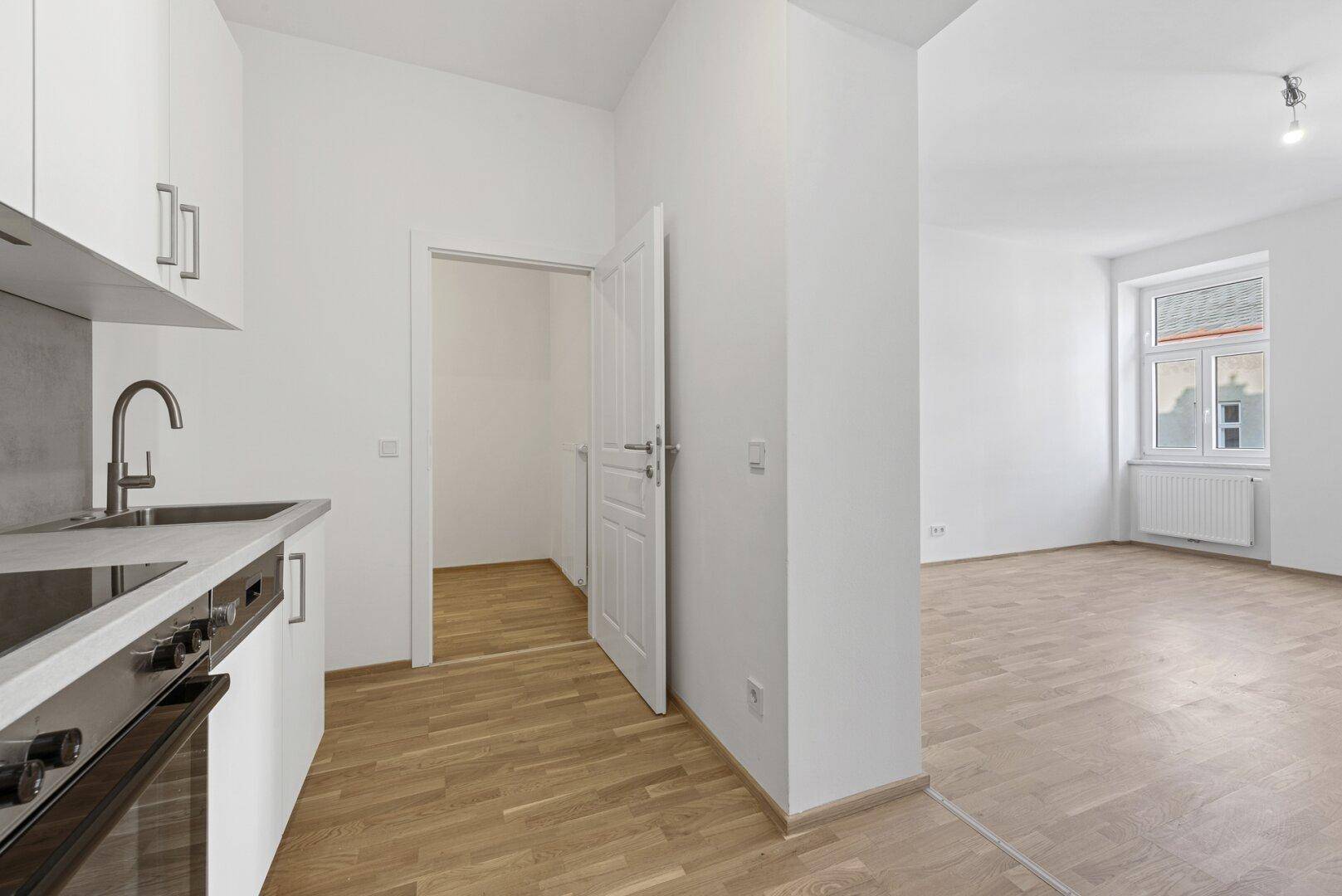 Heller Küchenbereich mit Holzboden und offenem Zugang zu einem weiteren Zimmer mit Fenster.