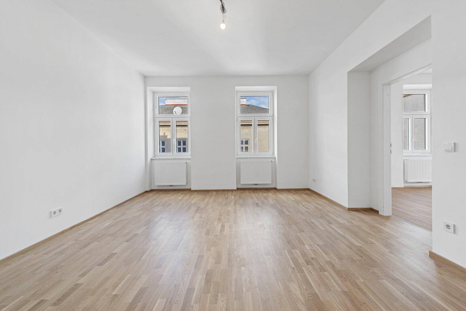 Geräumiges, helles Wohnzimmer mit zwei großen Fenstern, Holzboden und weiß gestrichenen Wänden.