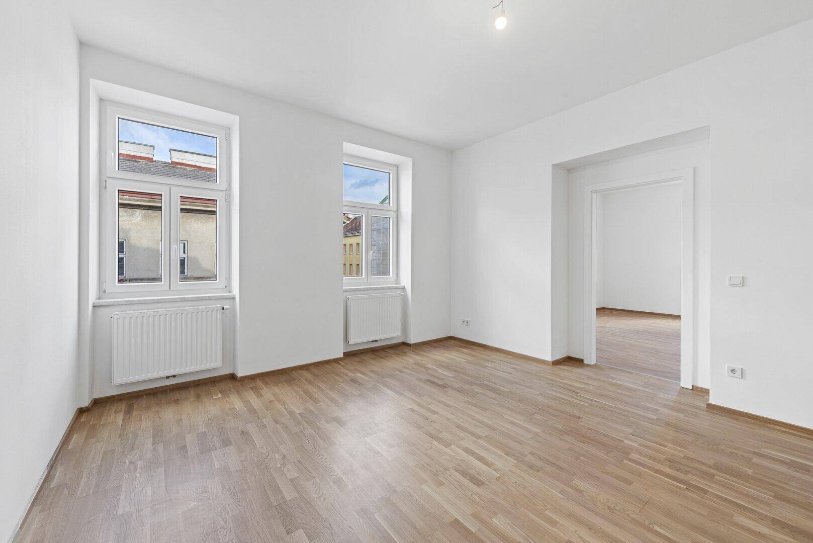 Heller Wohnbereich mit Holzboden, hohen Fenstern und Blick auf die umliegenden Gebäude.