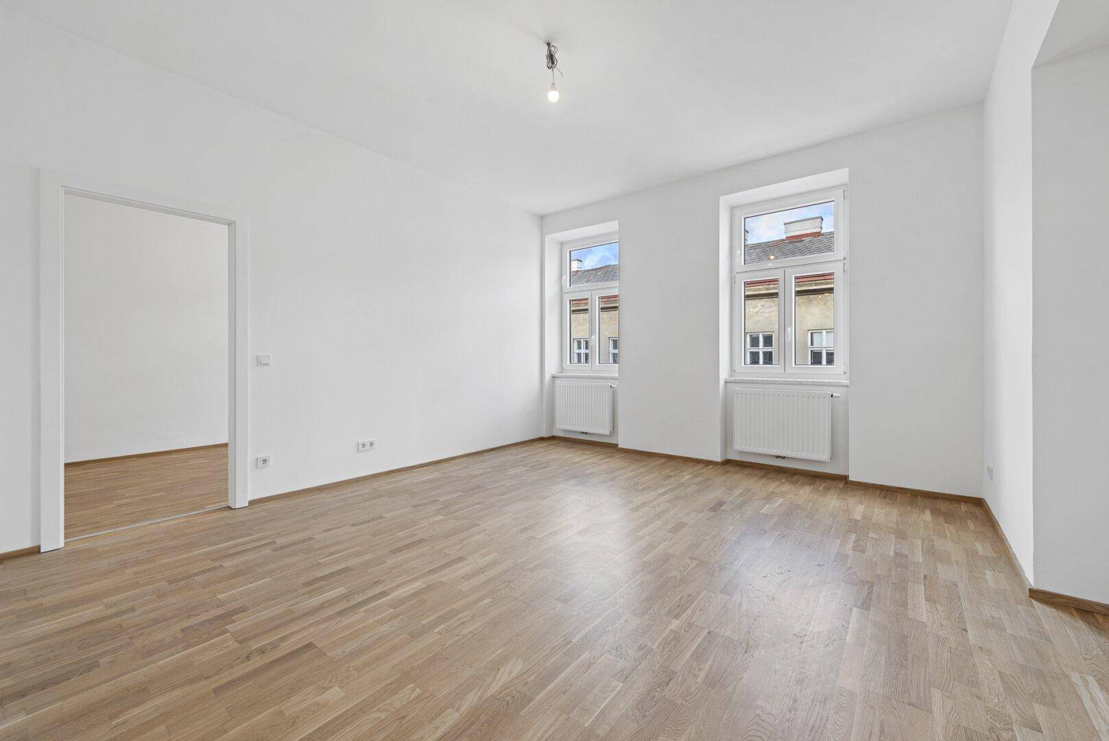 Lichtdurchfluteter Wohnraum mit Holzboden, zwei Fenstern und offenem Zugang zu einem weiteren Zimmer.
