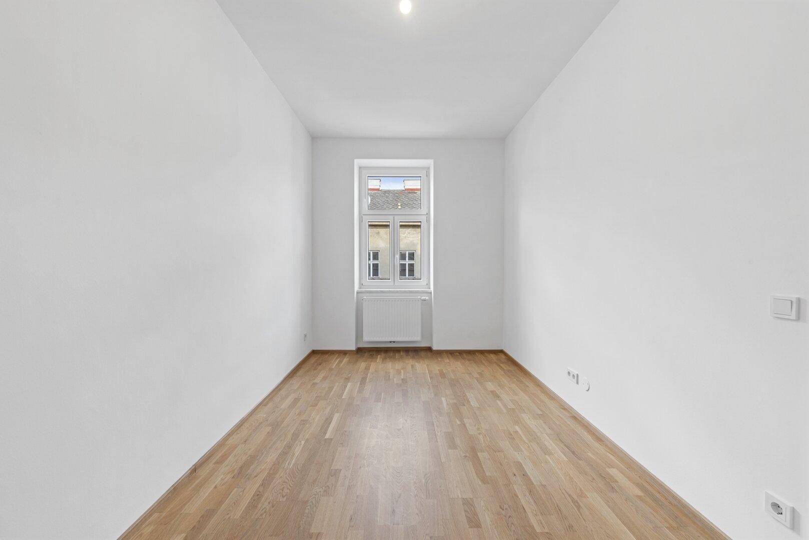 Schmaler, heller Raum mit Holzboden und einem Fenster, ideal als Schlaf- oder Arbeitszimmer.