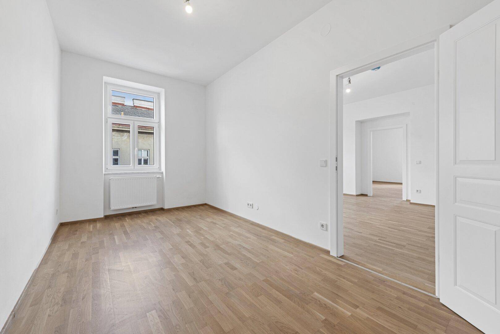 Helles Zimmer mit Holzboden und Fenster, bietet Zugang zu einem weiteren Wohnbereich.
