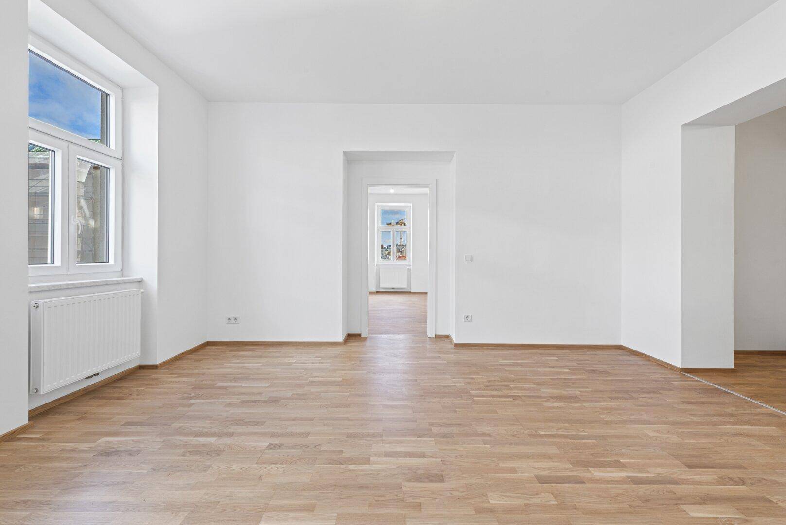 Geräumiger Wohnraum mit Holzboden, großen Fenstern und offenem Durchgang zu angrenzenden Zimmern.
