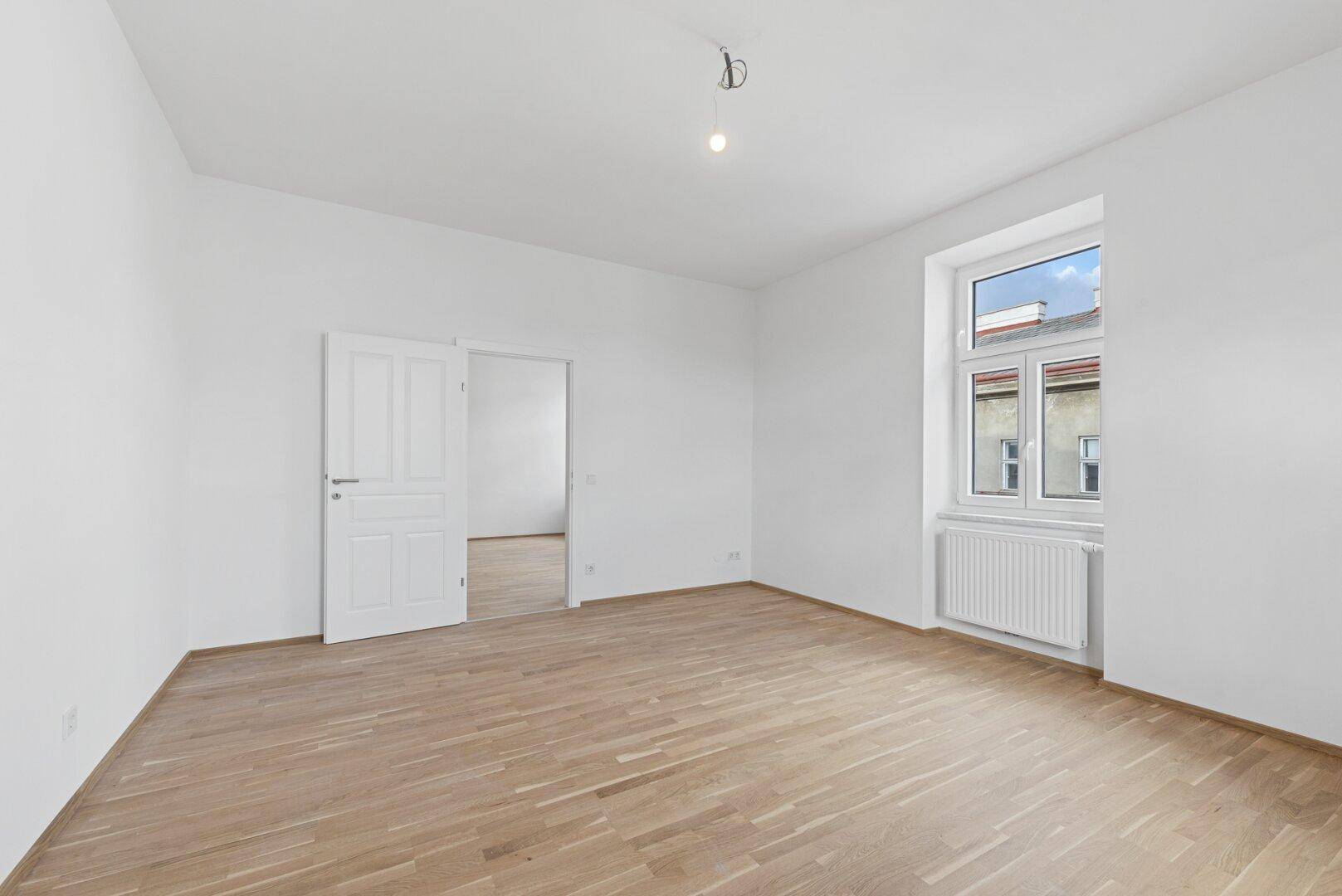 Helles Zimmer mit Holzboden und einem Fenster, bietet Zugang zu einem weiteren Raum.