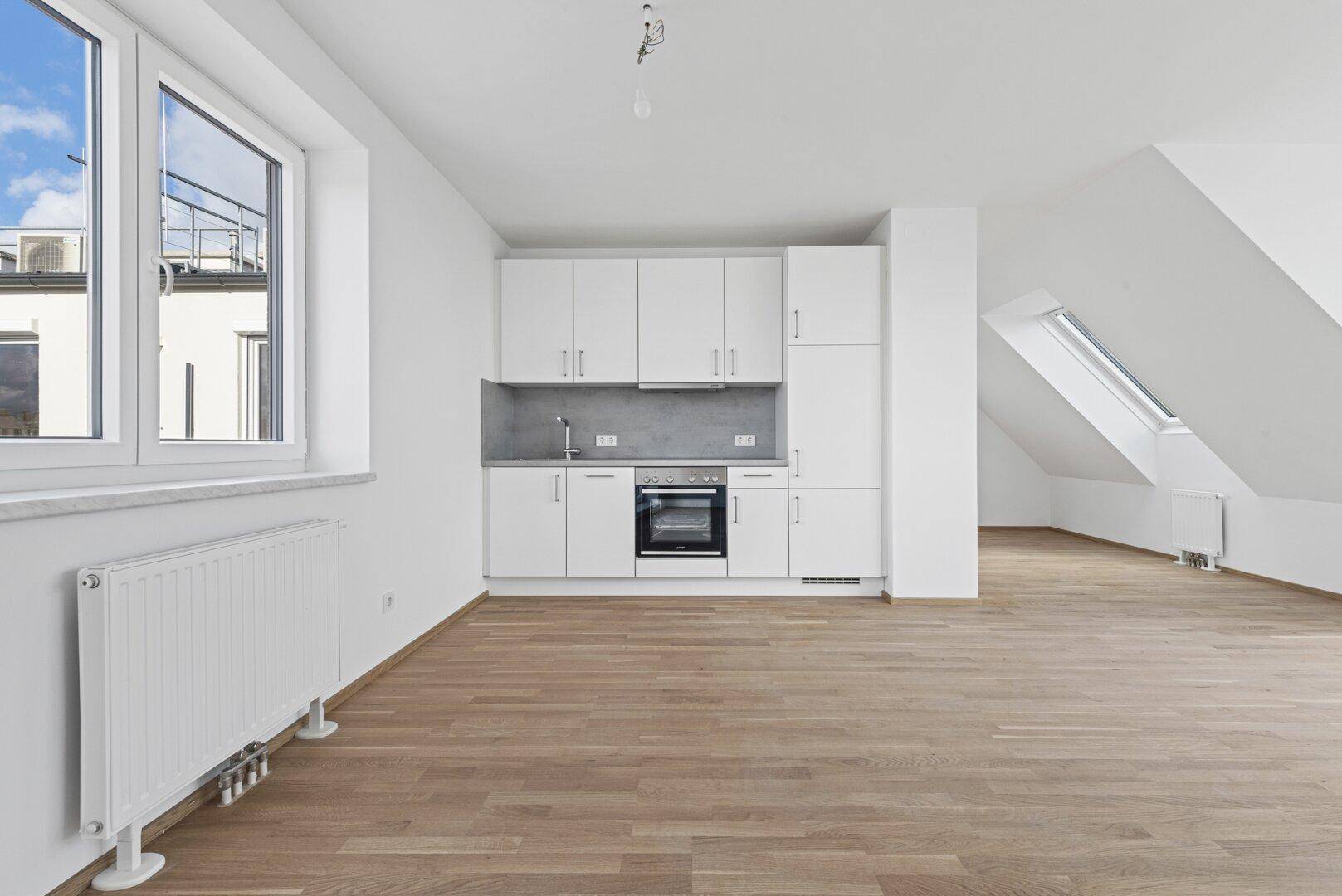 Offener Wohnbereich mit integrierter weißer Küchenzeile, Holzboden und Dachschräge.