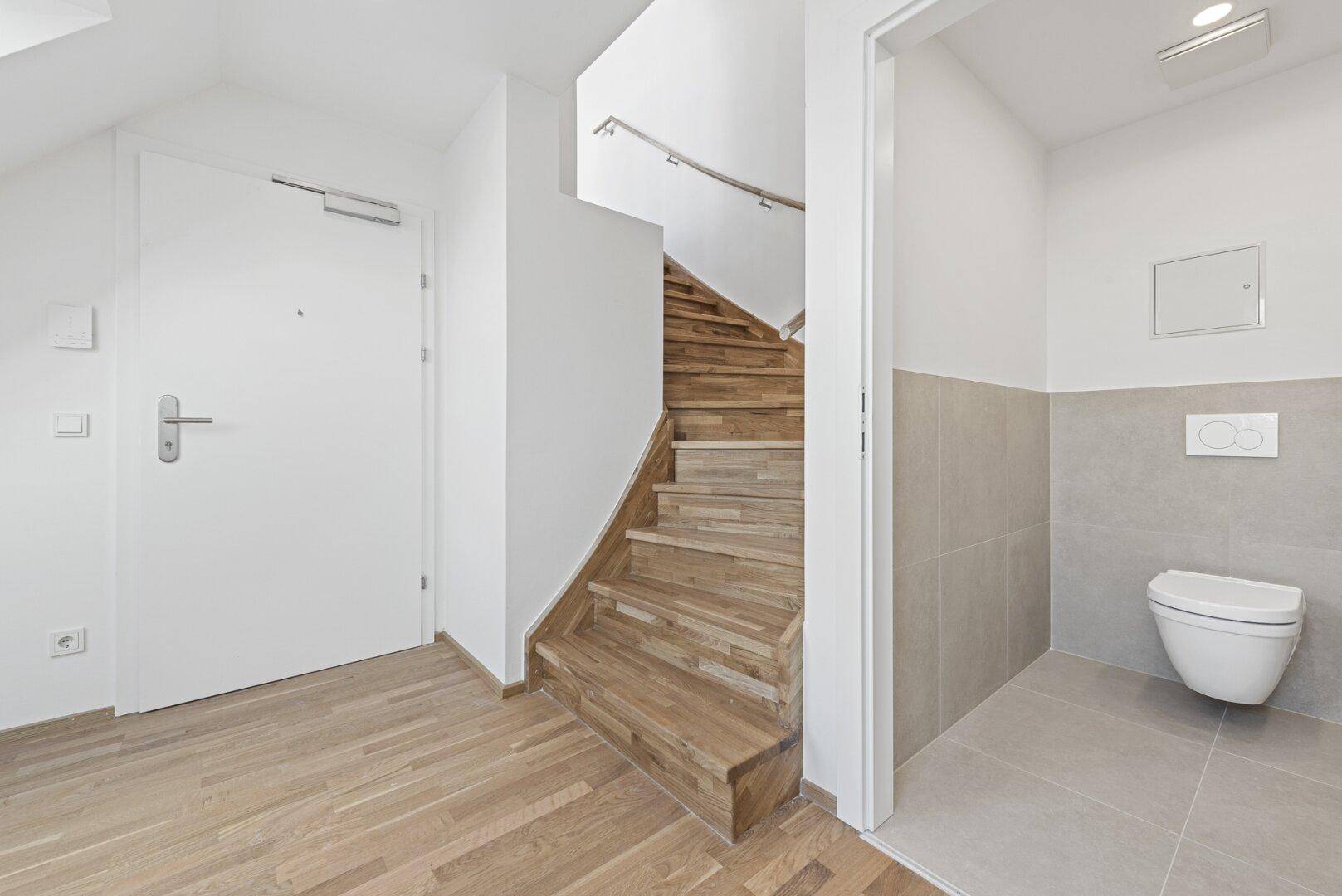 Heller Flurbereich mit Holztreppe, Eingangstür und Zugang zu einem modernen Gäste-WC.